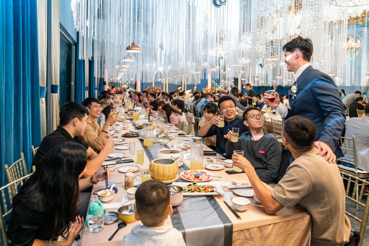 Guests seated at long dining tables in a modern restaurant with shimmering metallic curtains and elegant decor.