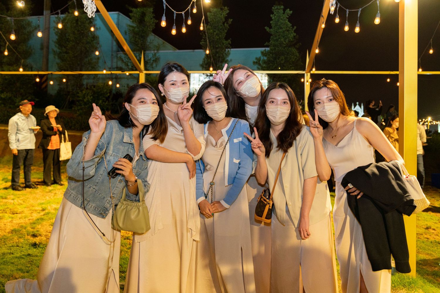 A group wearing protective face masks and matching beige dresses poses together at an outdoor evening event.