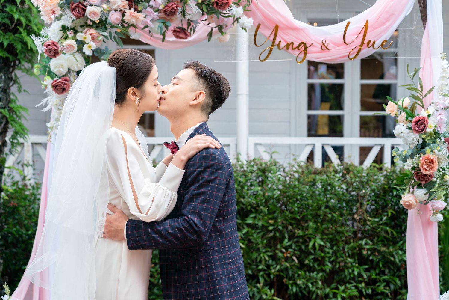 A romantic wedding kiss under an arch decorated with pink and red roses and a beautiful garden backdrop.