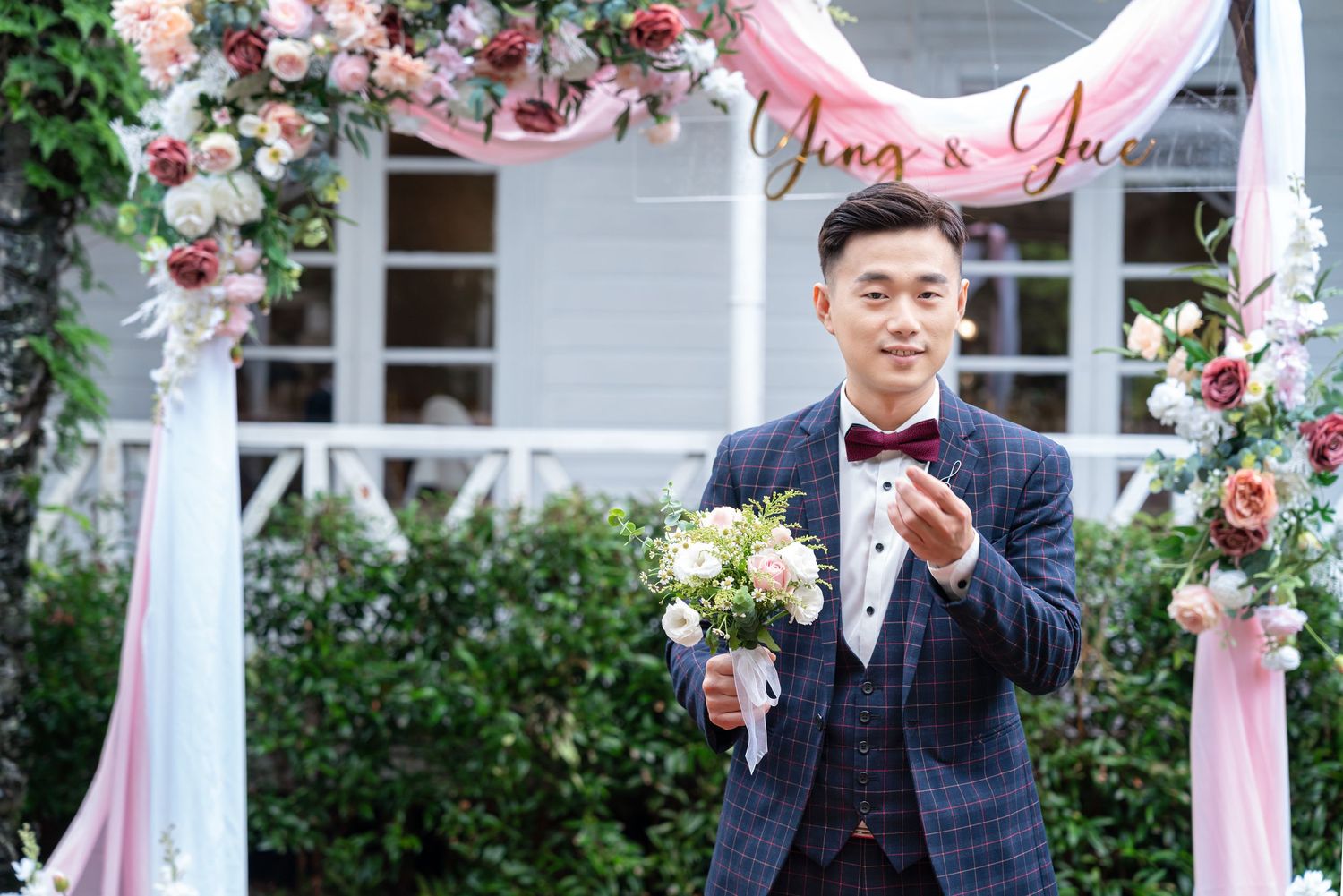 Wedding ceremony setting with a pink and floral decorated arch and groom in navy plaid suit holding a bouquet.