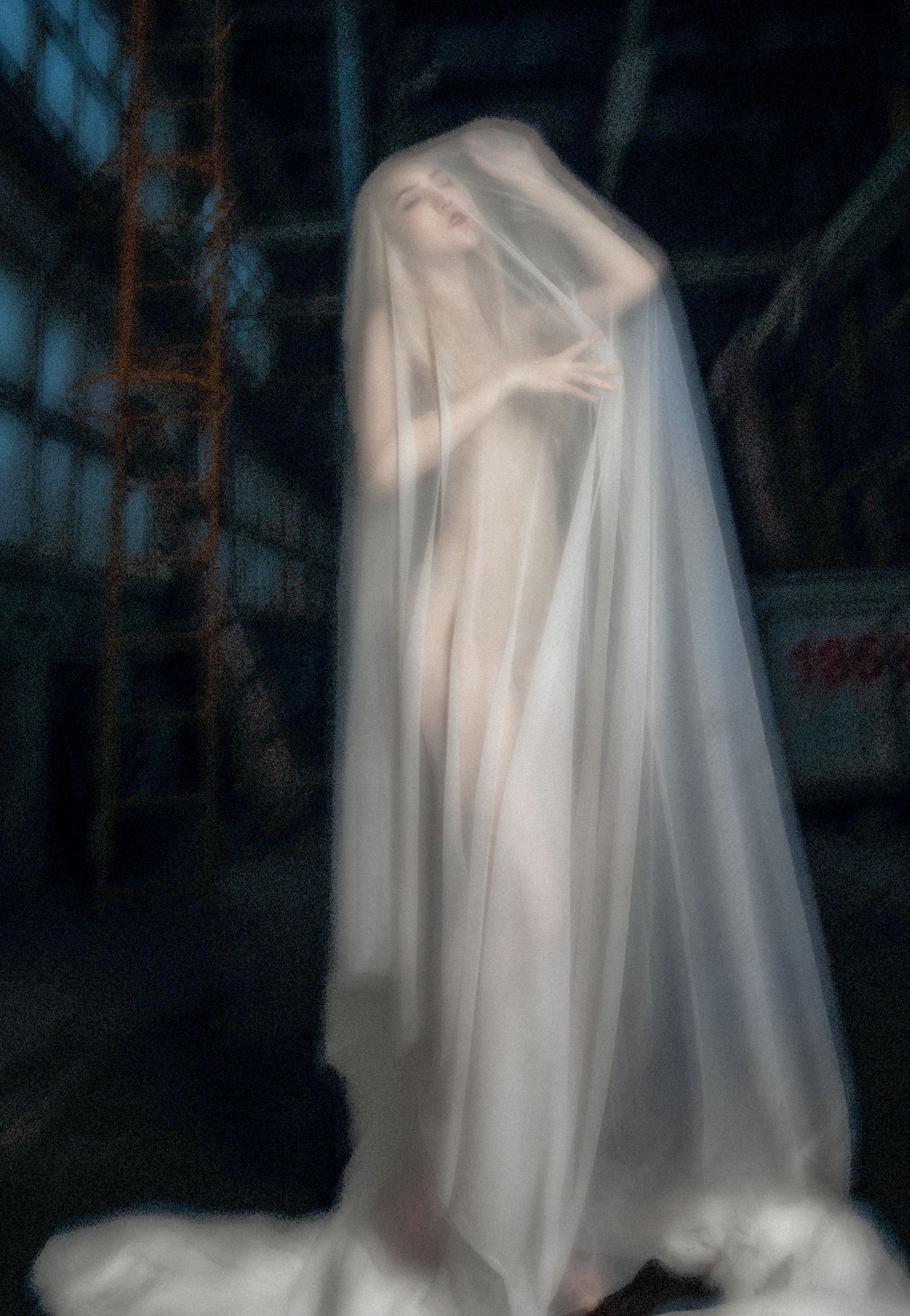 Ethereal figure in flowing white fabric poses dramatically in dark industrial space creating ghostly artistic effect.
