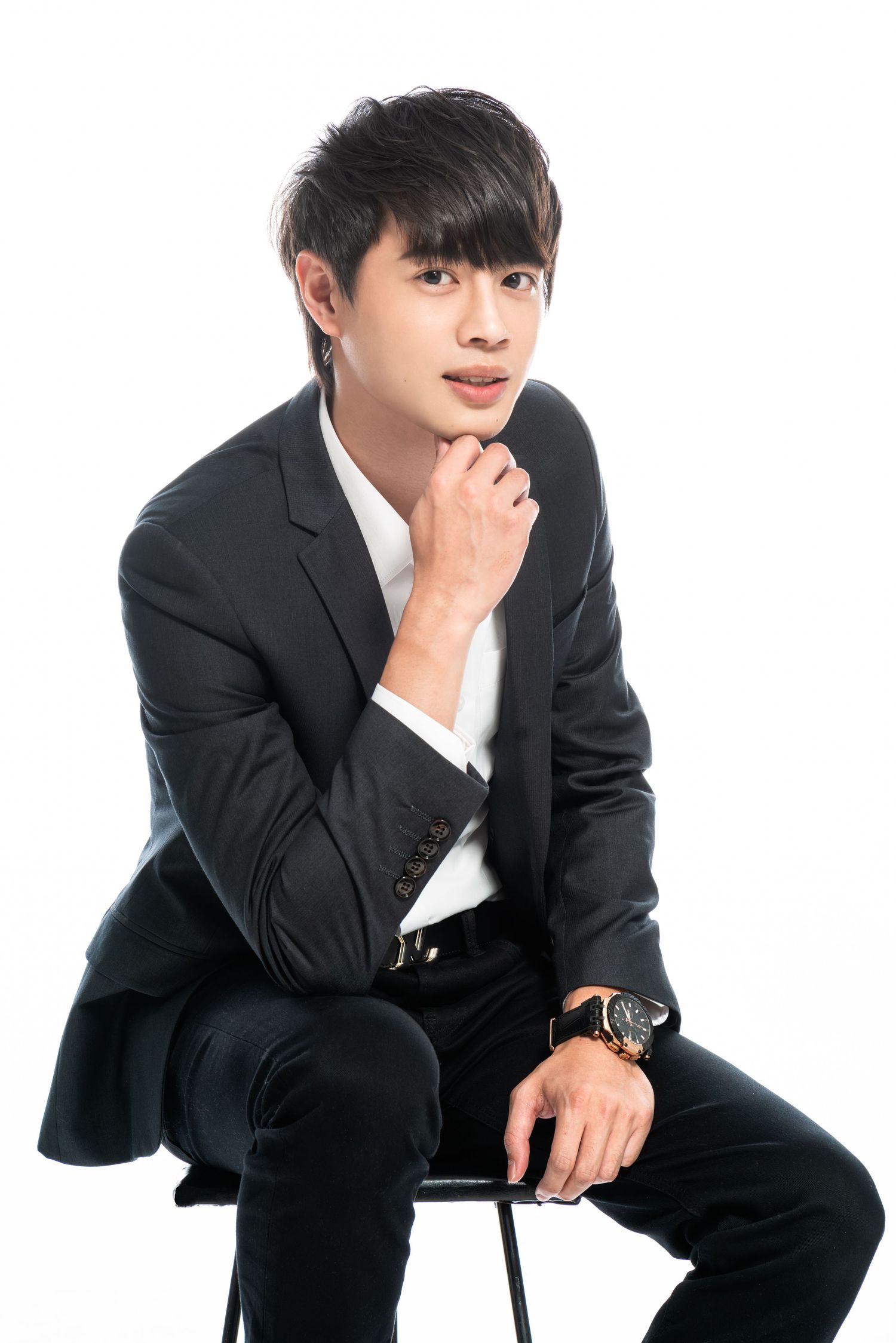 Young man in black suit and white shirt poses thoughtfully on chair against white background with wristwatch.