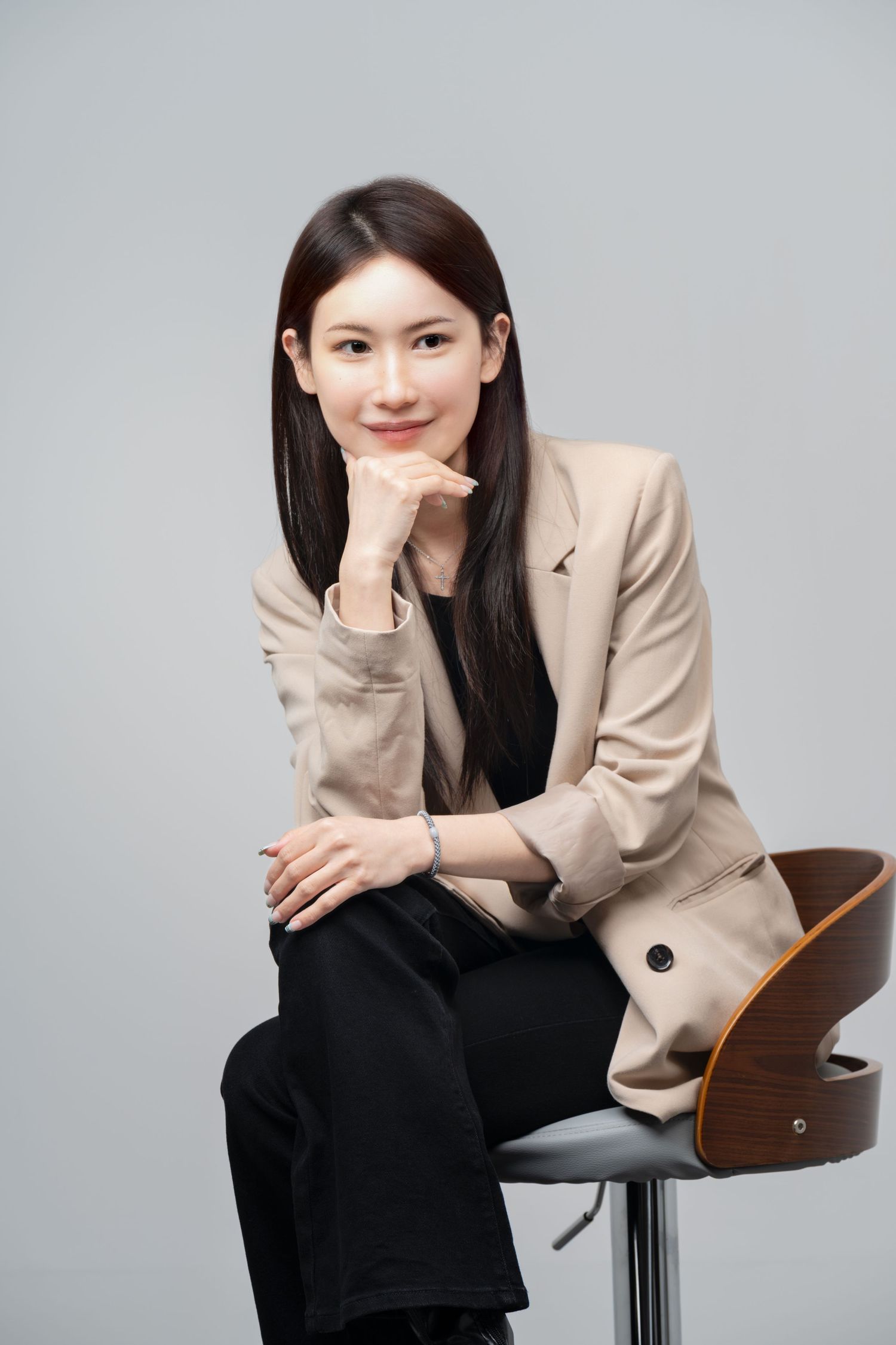 Professional portrait of someone in a beige blazer and black pants sitting on a modern wooden bar stool against gray background.