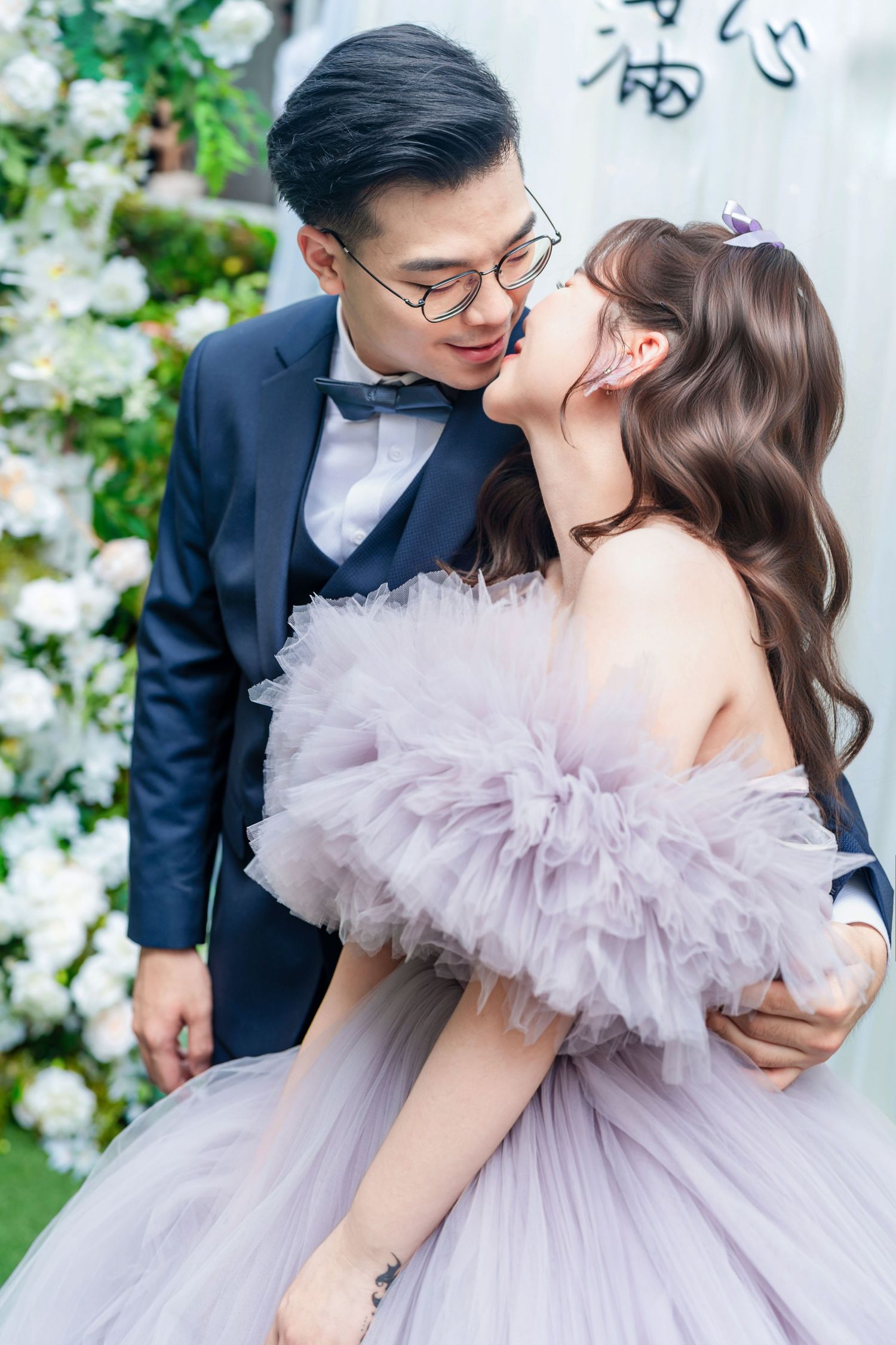 A romantic wedding photo with a bride in a lavender tulle gown kissing next to white floral decorations.