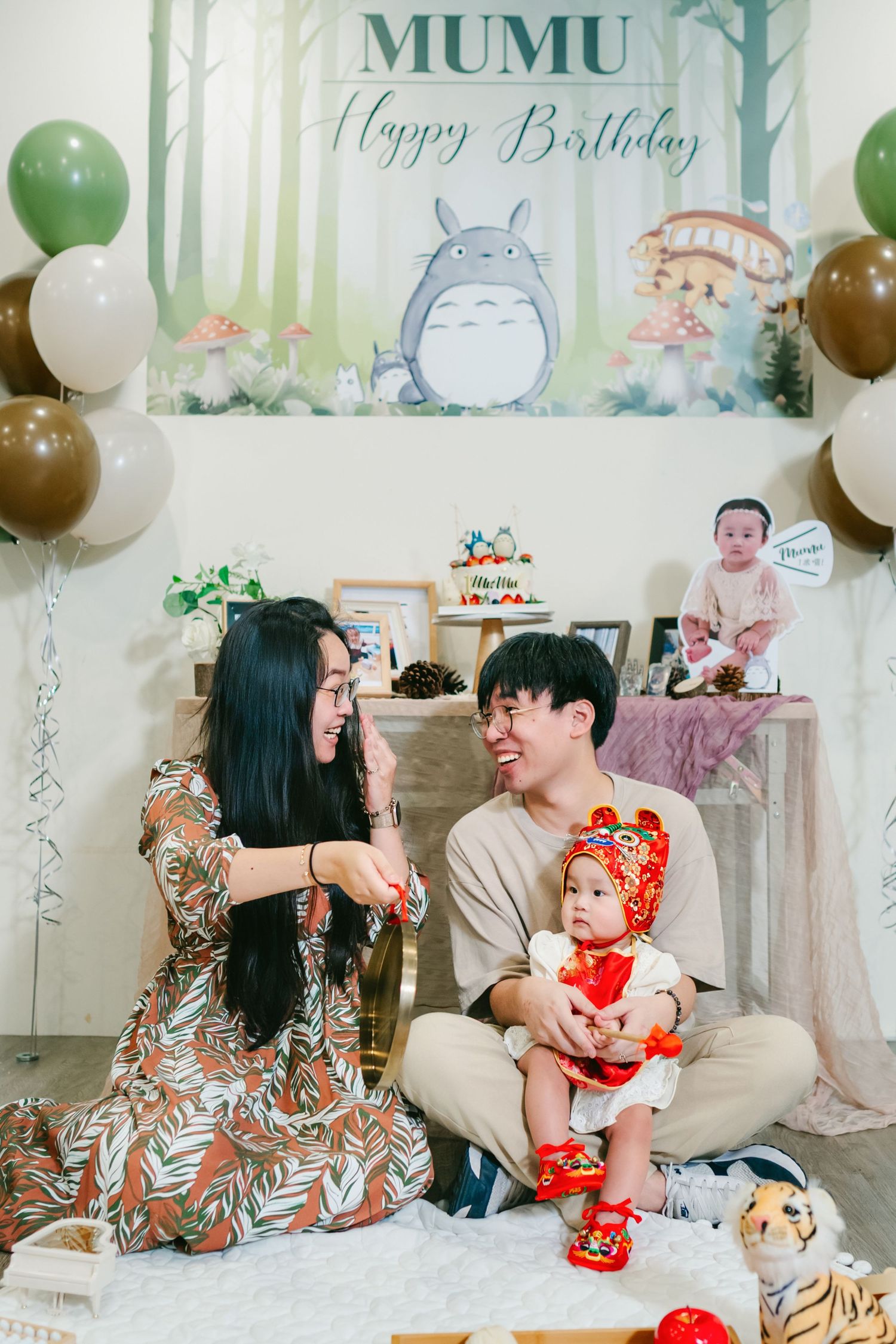 Birthday celebration with Totoro themed decorations, green and brown balloons, and baby in a red outfit on white couch.
