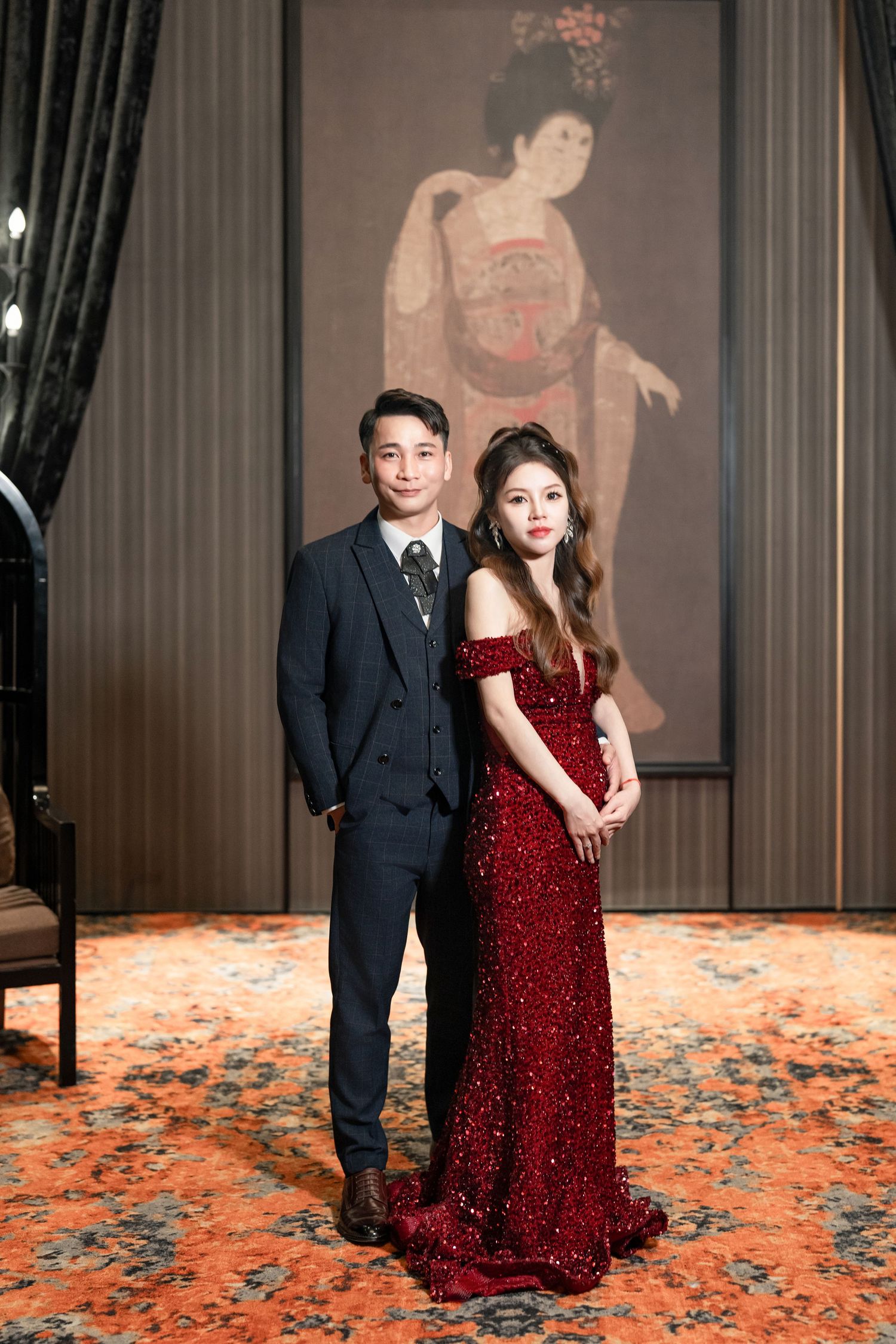 A couple poses together in formal attire, with one person wearing a sparkling red gown and another in a dark suit.