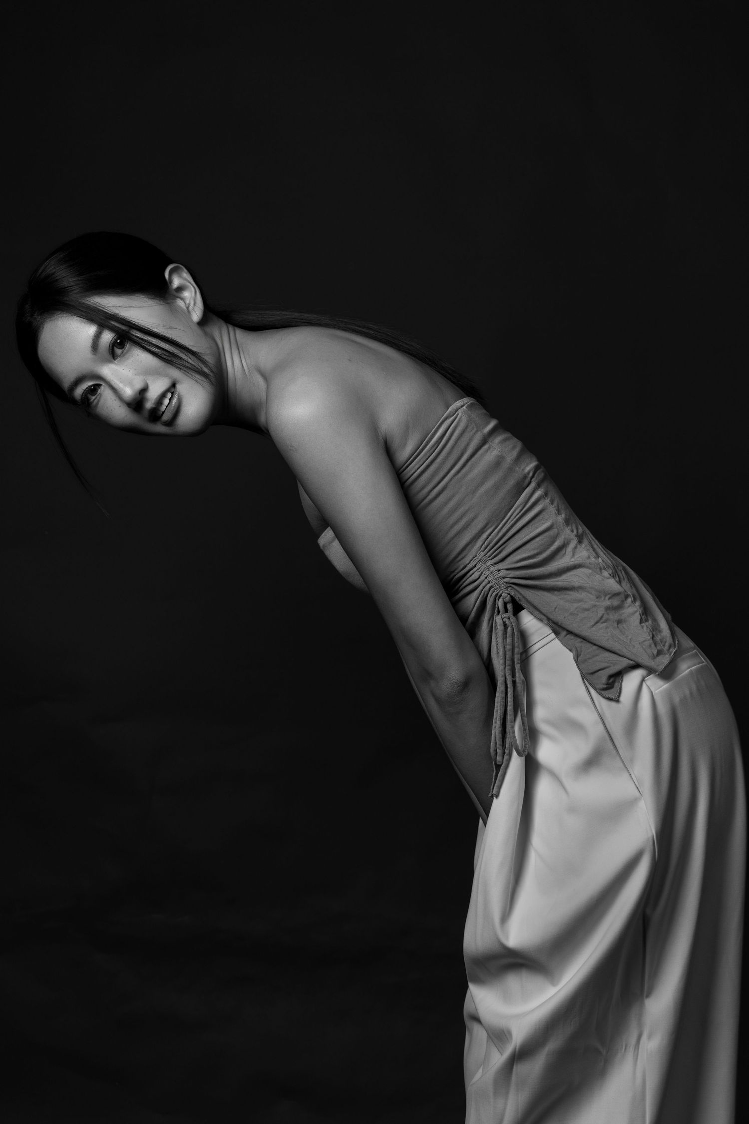 Black and white portrait of a woman in an elegant halter dress, leaning forward gracefully against a dark background.
