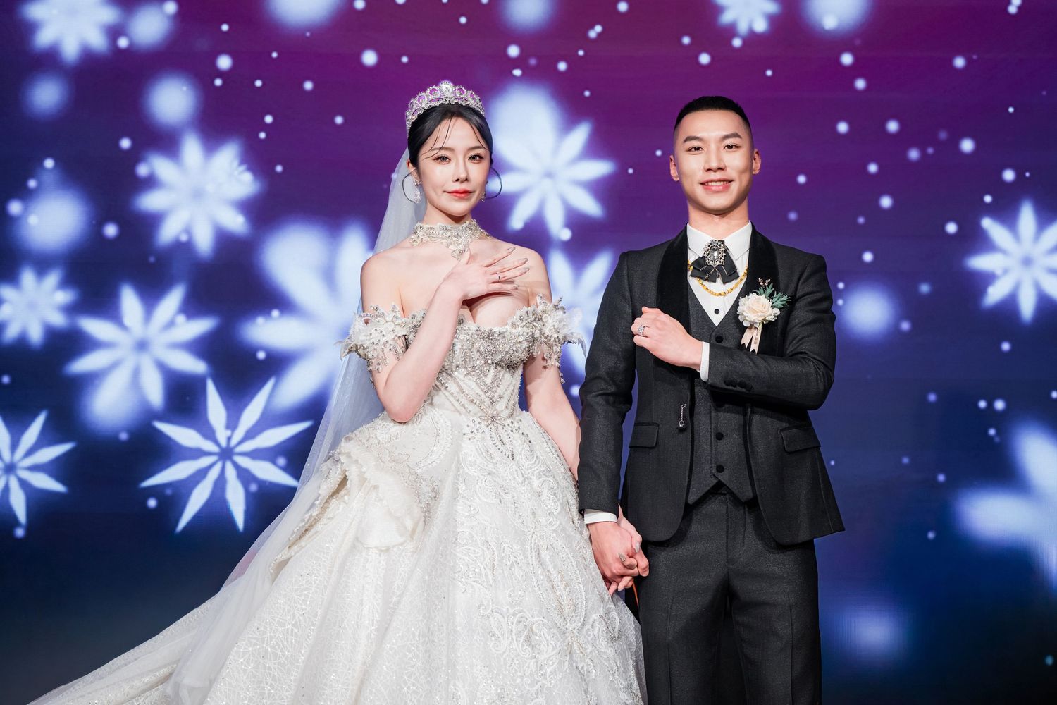 A couple in wedding attire poses on stage with blue snowflake projections and lighting effects in the background.