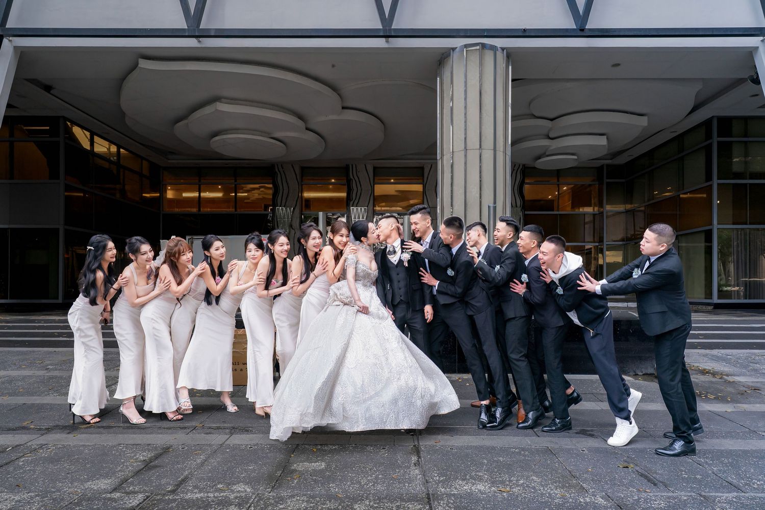 Wedding party poses creatively along a modern architectural building with glass and metal features.