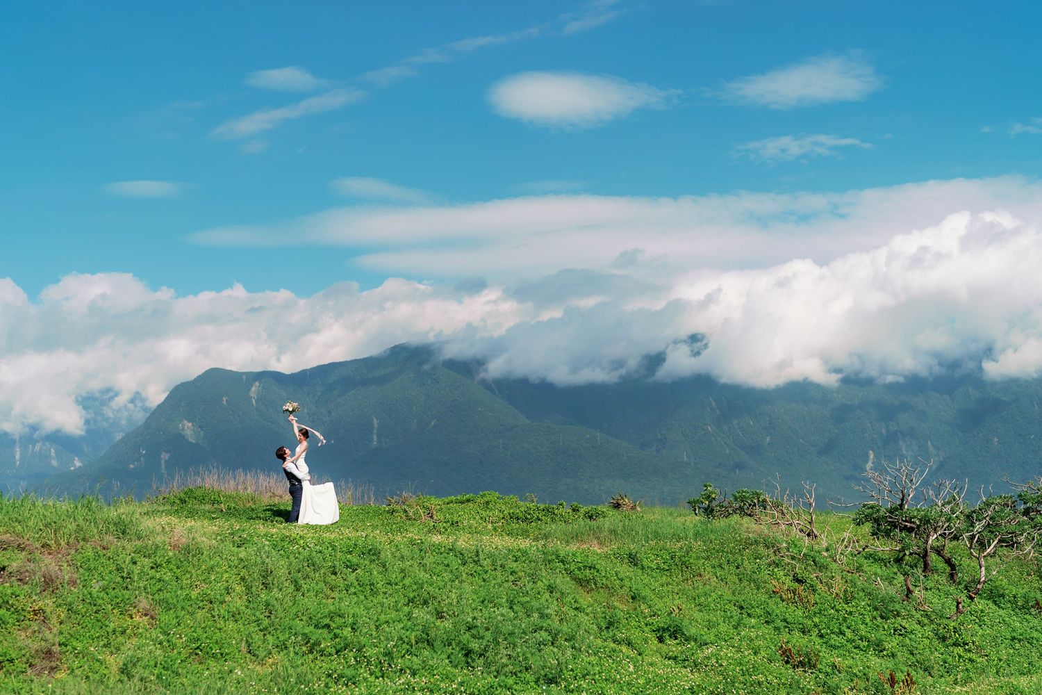 A lone figure in white stands on a green hilltop against a backdrop of majestic mountains and blue sky with clouds.