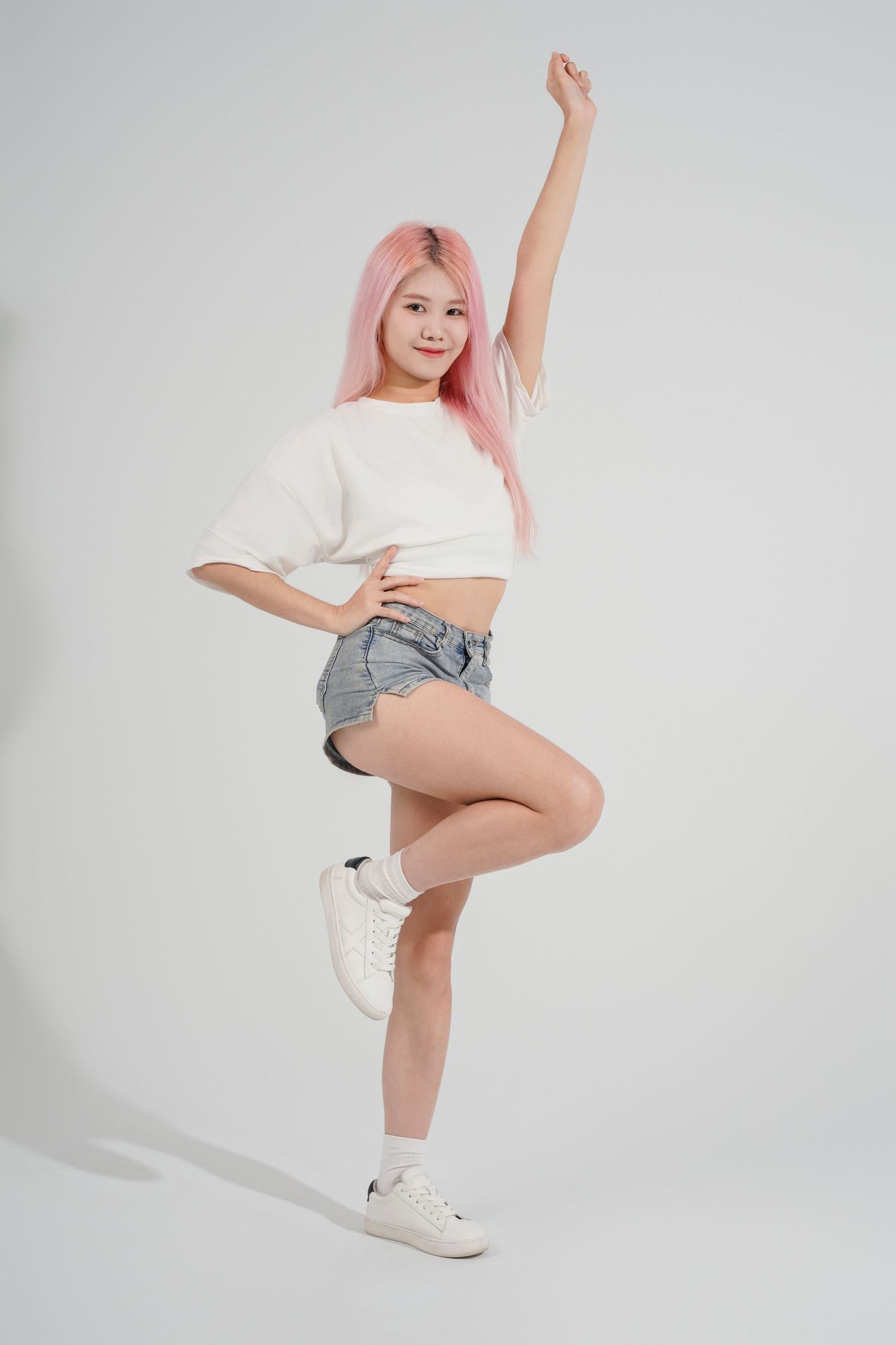 Person with pink hair in white crop top and denim shorts striking energetic dance pose against light background.