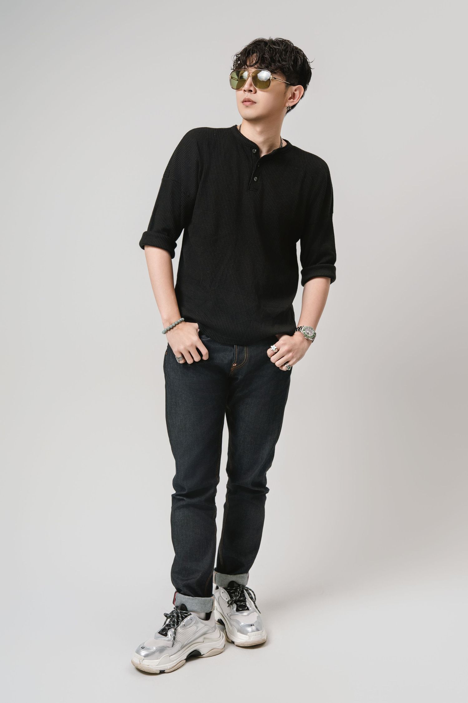Person wearing all black casual outfit with white and gray sneakers poses against light gray studio background.