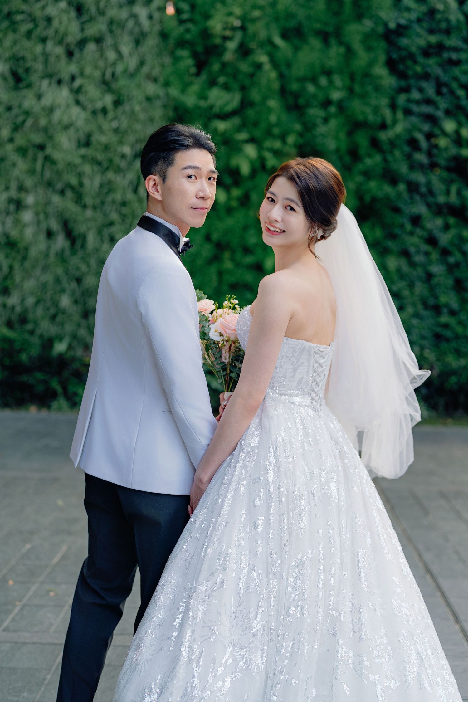 A bride and groom pose together outdoors, with the bride in a white ballgown and the groom in a white tuxedo jacket.