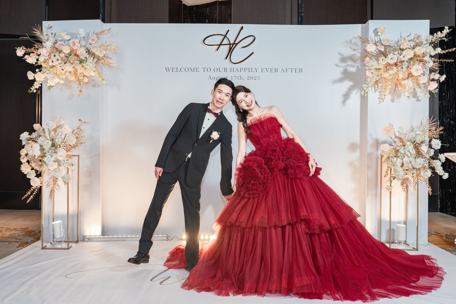 Couple in wedding attire poses elegantly against light backdrop with floral decor and lanterns in romantic winter setting.