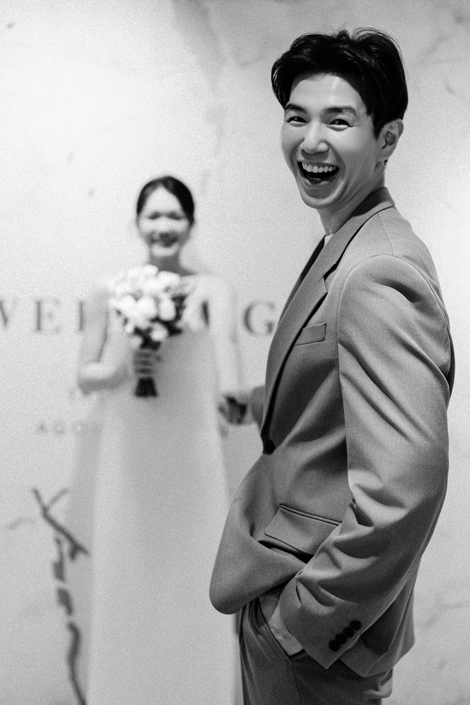 Black and white wedding photo showing a joyful moment with the groom laughing while the bride holds flowers in background.