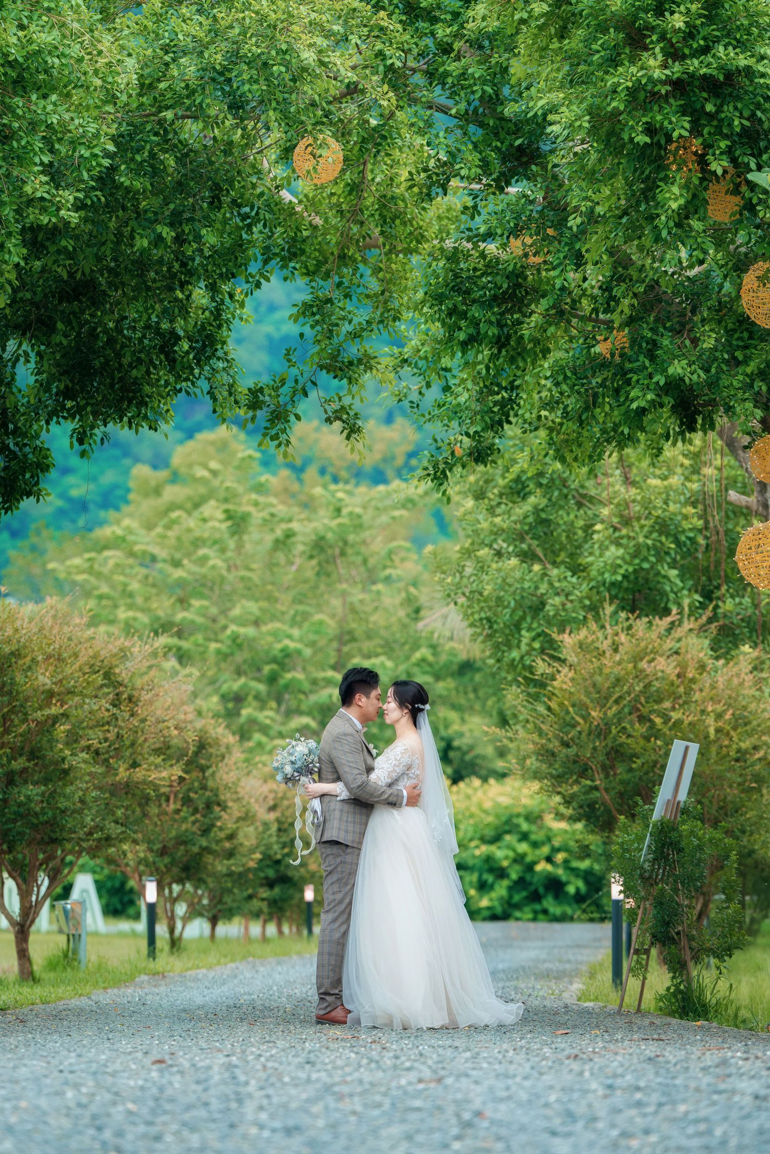 A couple wearing wedding attire share a romantic moment while sitting on a stone path surrounded by lush greenery.