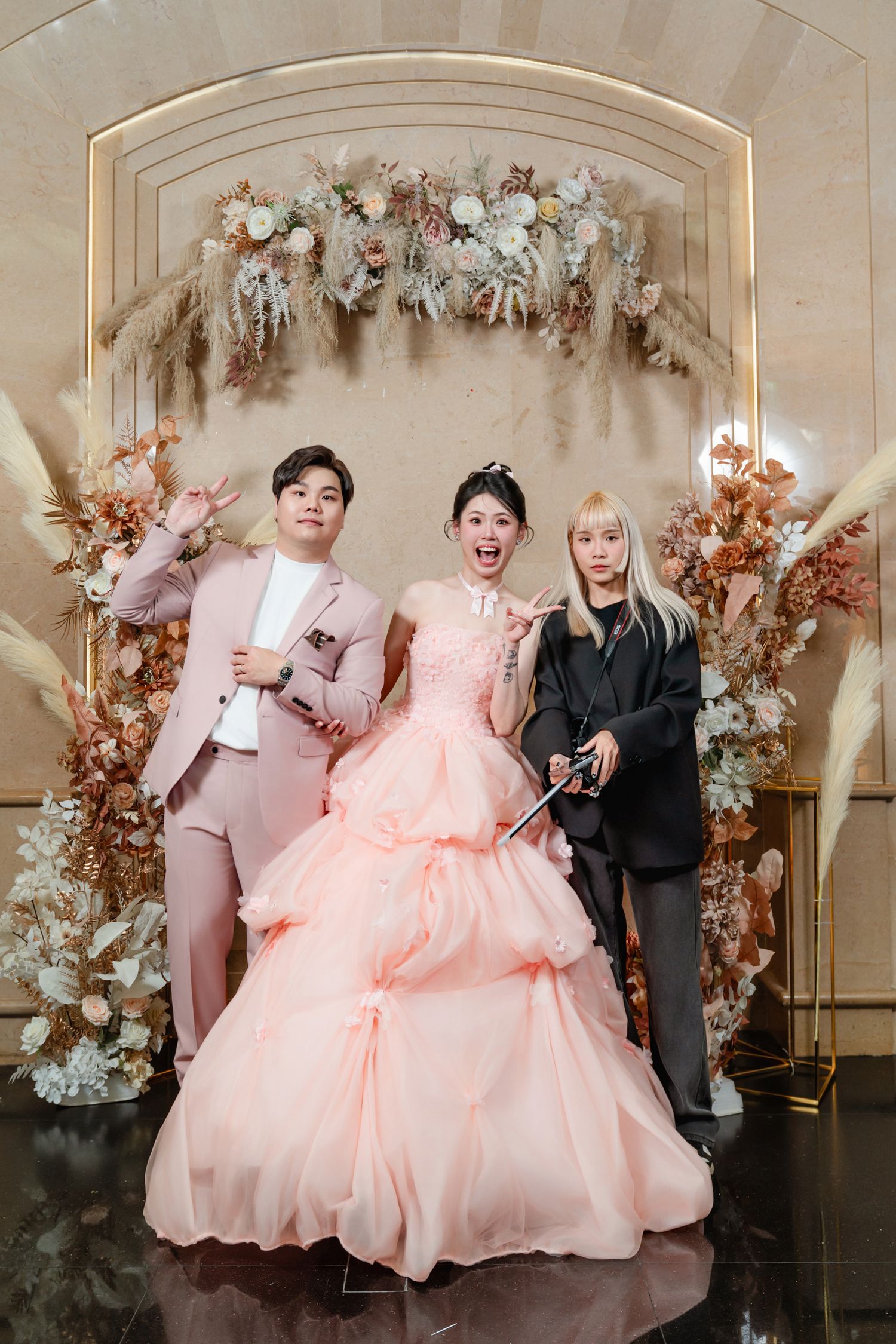 An elegant wedding celebration under a floral arch with three people in formal attire including a pink ballgown.