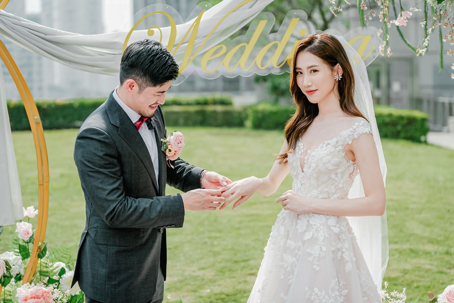 A couple at their outdoor garden wedding ceremony with gold decor and floral arrangements as the groom puts a ring on his bride.