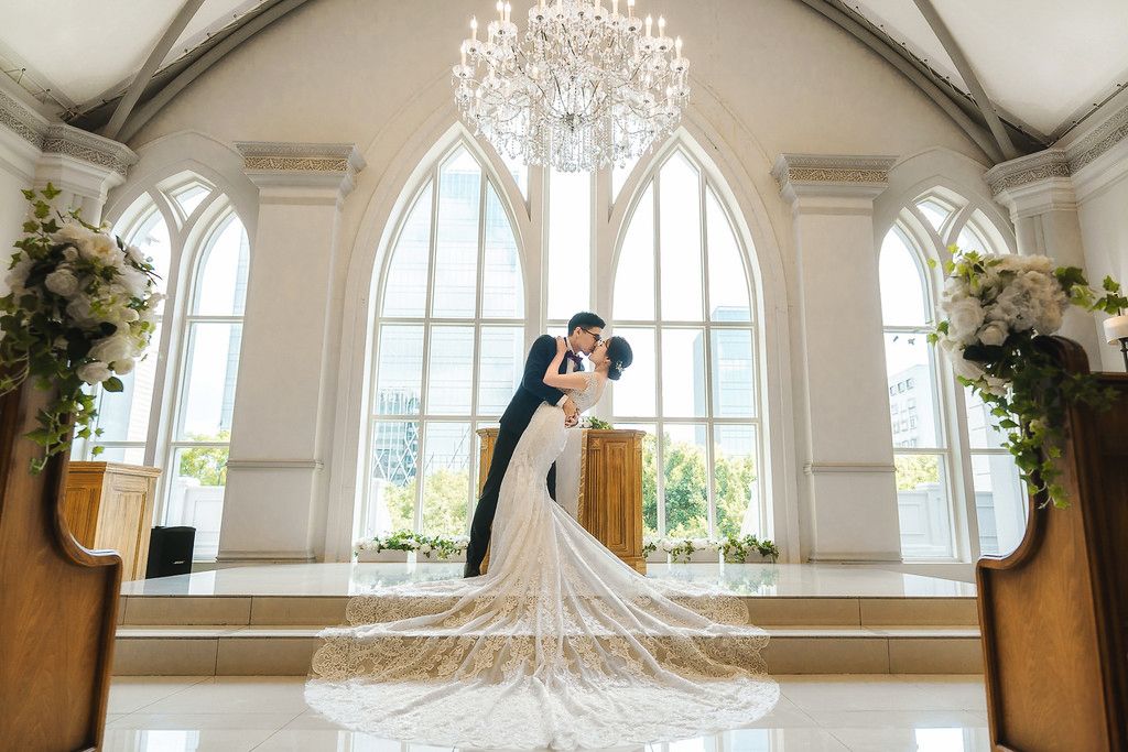 A couple embraces in an elegant white chapel with arched windows, tall pillars and a grand crystal chandelier overhead.