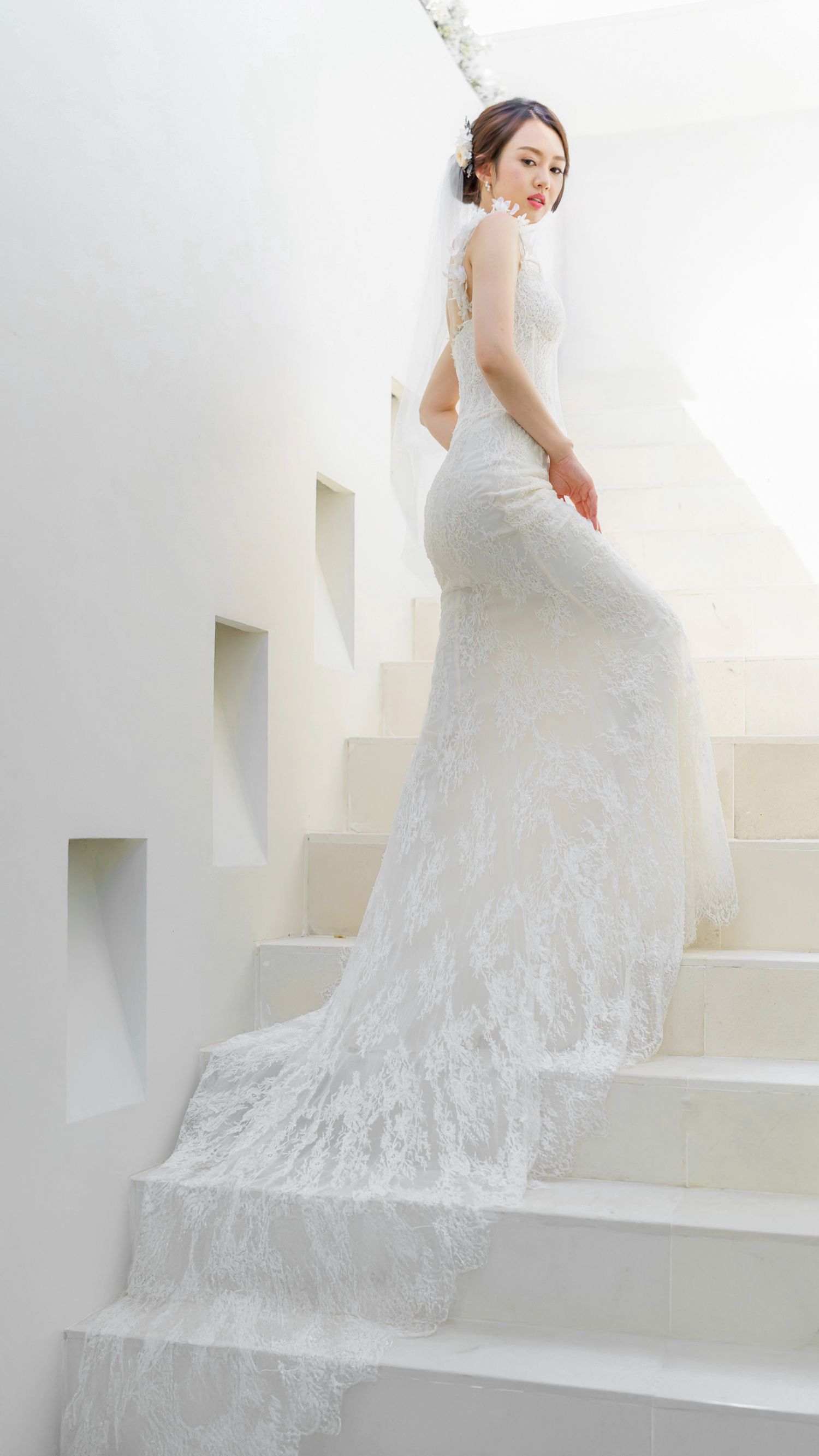 A bride in an elegant lace wedding gown with flowing train poses gracefully on white stairs in a bright setting.