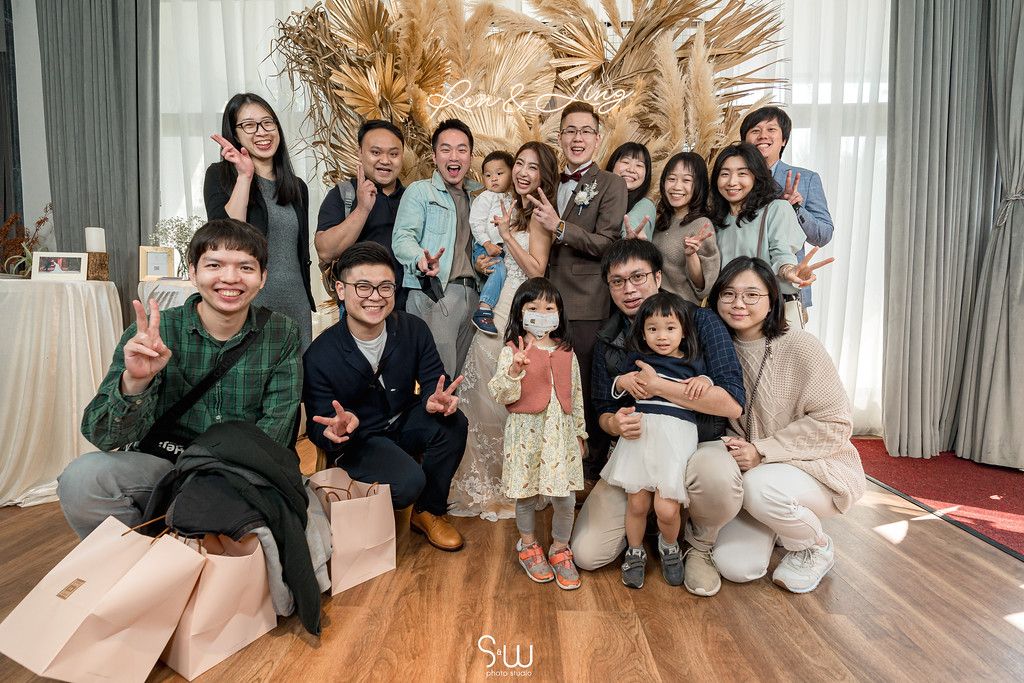 A large group gathers indoors for a casual celebration photo with dried pampas grass decor in the background.
