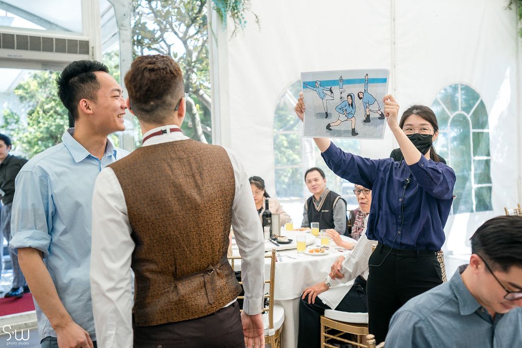 A group celebrates at a wedding reception in a light-filled tent with hanging greenery.