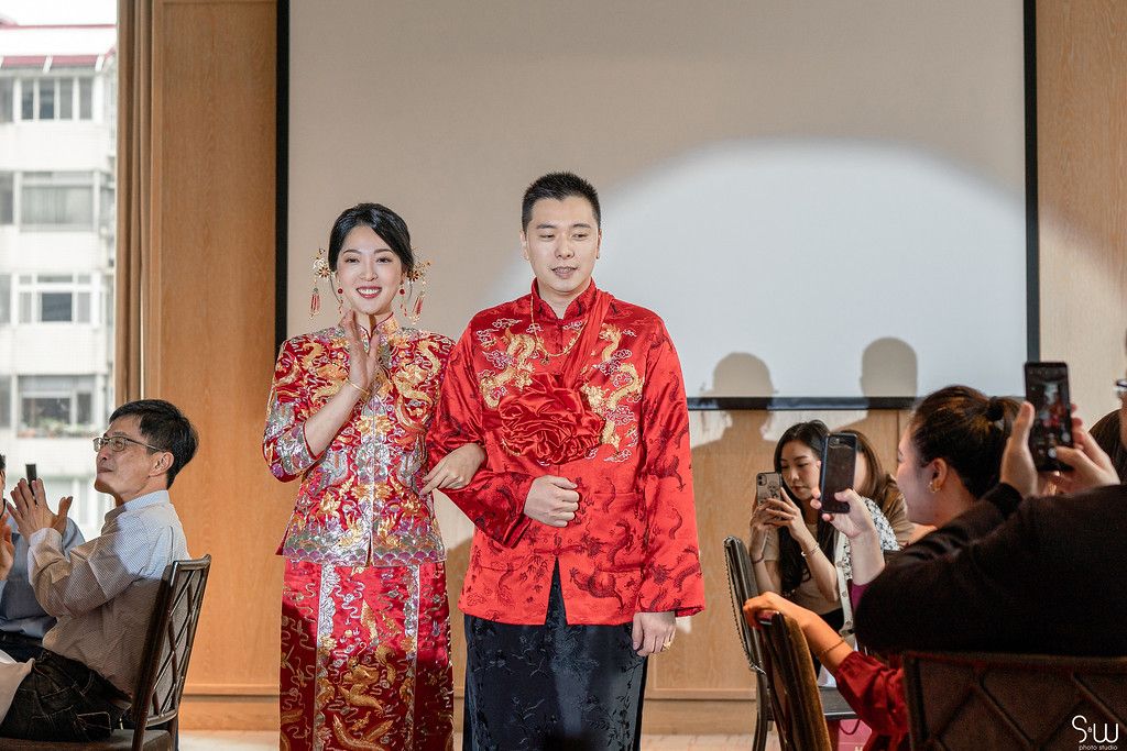 A couple in red traditional Chinese wedding attire stand on stage while guests take photos.