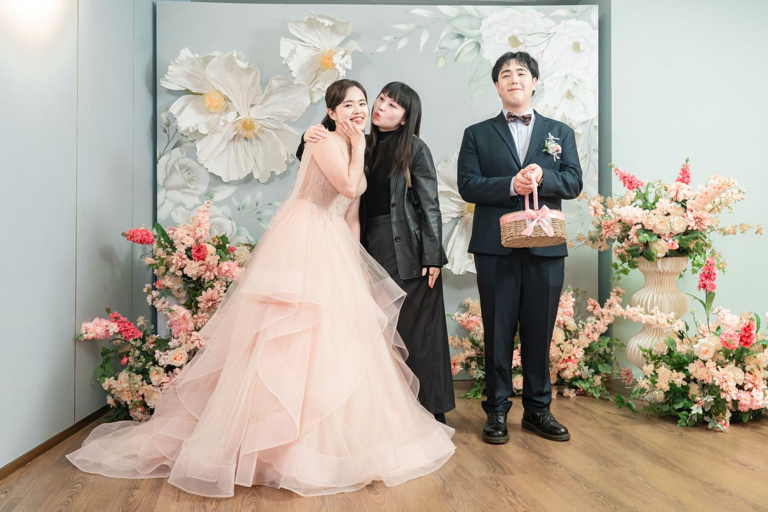 A group celebrates in formal wear with pink dresses and suits against a floral backdrop during an event.
