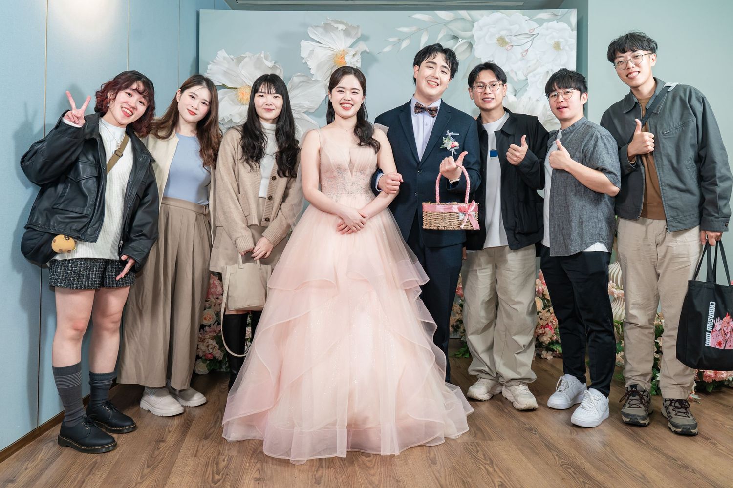 A group of people gather together for a formal event wearing a mix of suits and elegant dresses in pink and neutral tones.