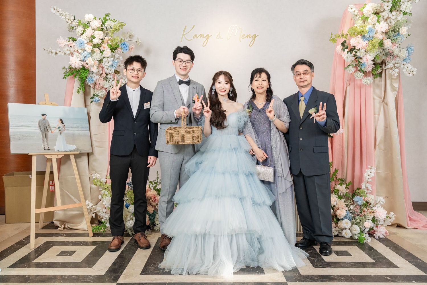 Wedding party poses together in elegant formal attire with floral decorations and soft pink backdrop.