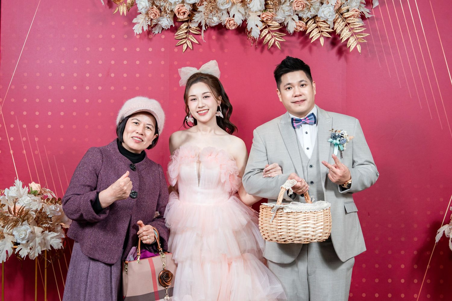 Three people pose against a pink polka dot backdrop with floral decorations, wearing formal attire and holding baskets.