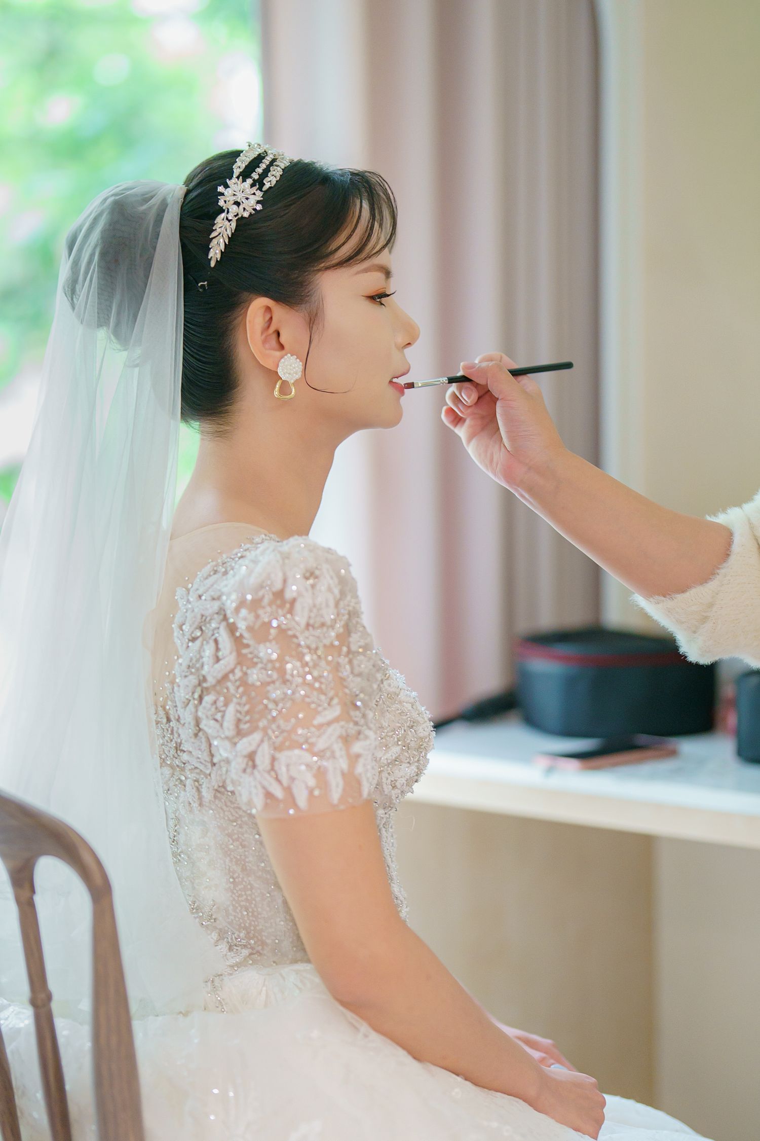 A bride in a delicate lace wedding dress with sheer details gets her makeup done during wedding preparations.
