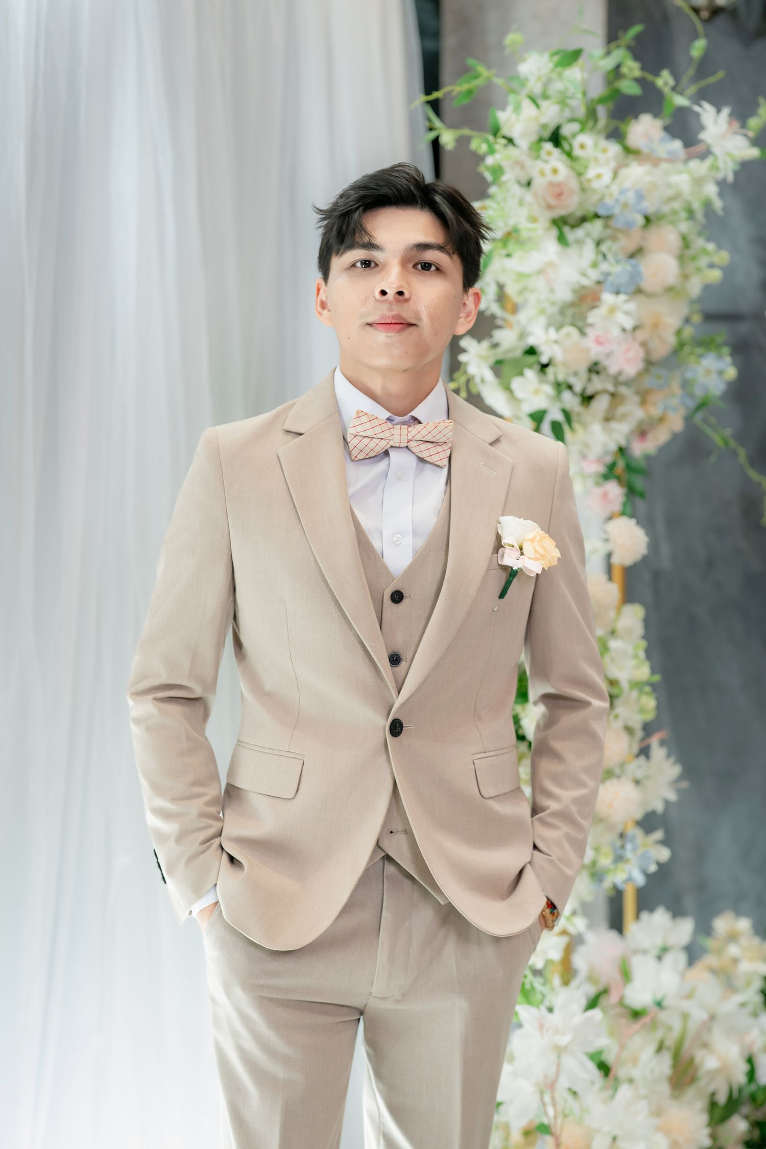 Elegant groom in beige three-piece suit with bow tie and boutonniere poses before floral wedding backdrop.