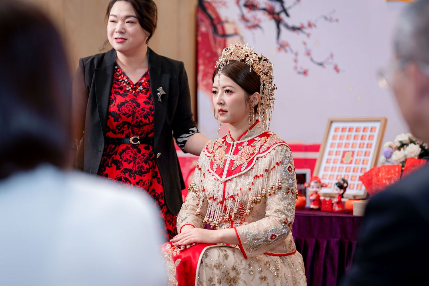 Bride in an ornate gold and red traditional Chinese wedding gown and headdress sits while a smiling woman in a red dress stands beside her.