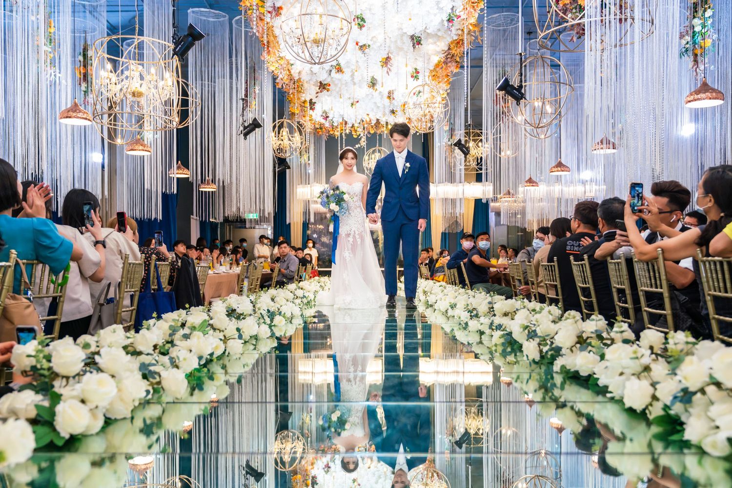 Elegant wedding ceremony in luxurious chapel with crystal chandeliers and white flower aisle arrangements along mirrored floor.