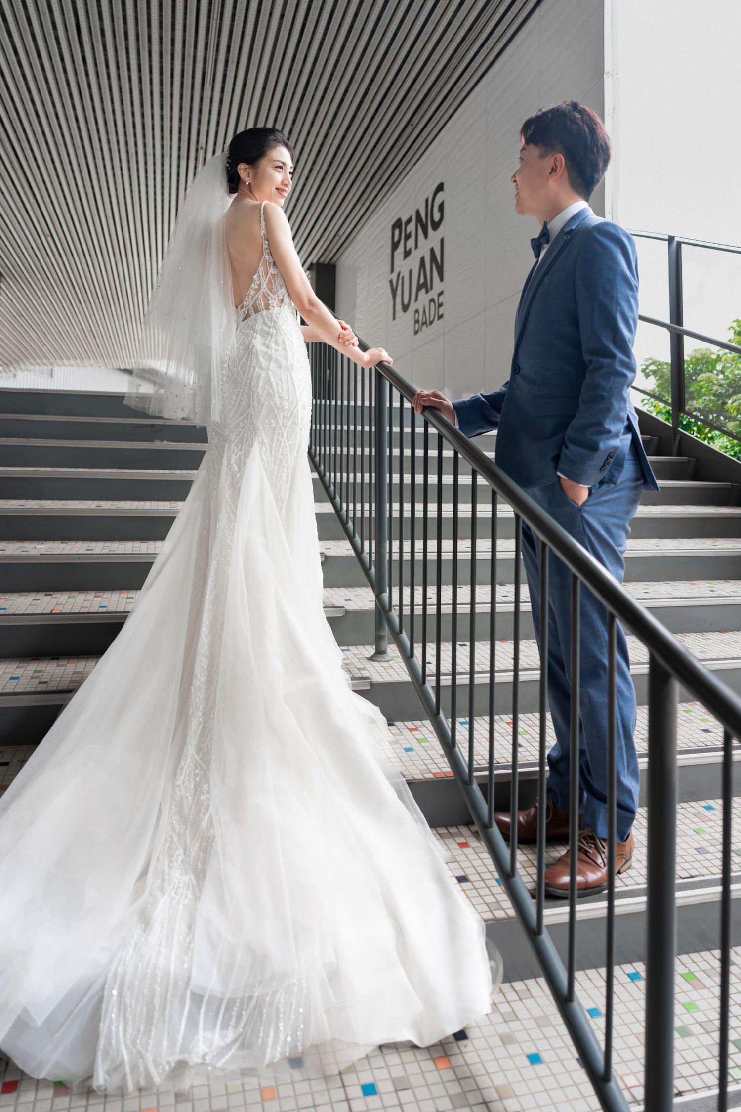 A bride in a flowing lace wedding gown stands on modern stairs while meeting someone in a navy suit.