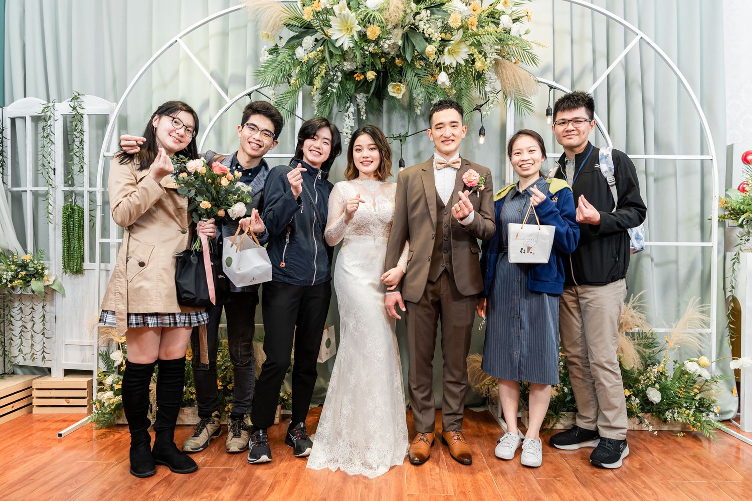 Group photo at a wedding celebration with floral arch backdrop and warm lighting.