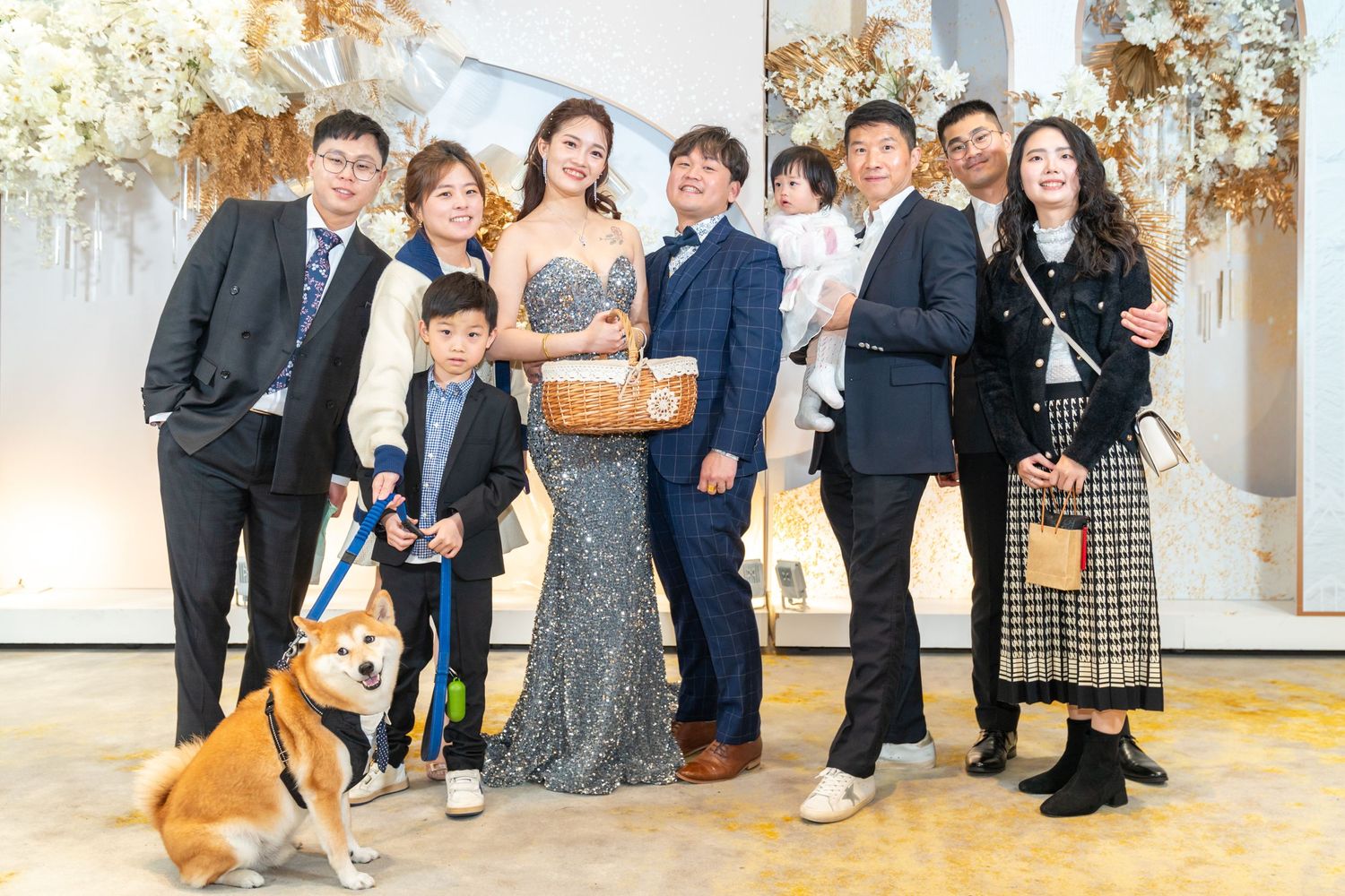 Group in formal attire poses together with a small orange dog against an elegant backdrop with floral decorations.