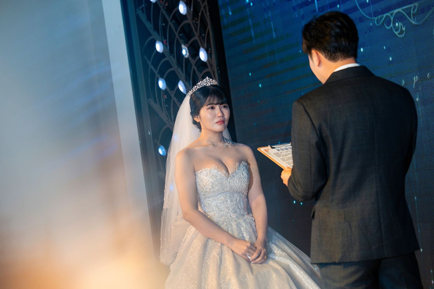 Bride in elegant beaded gown and tiara smiles at groom in black suit during wedding ceremony.