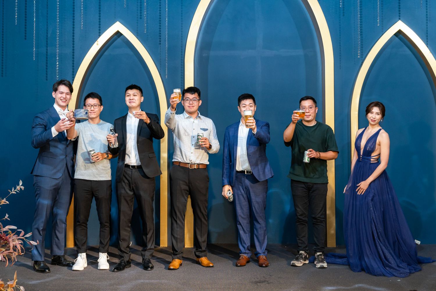 Group of people posing against a blue wall with white arched design wearing formal and casual attire.