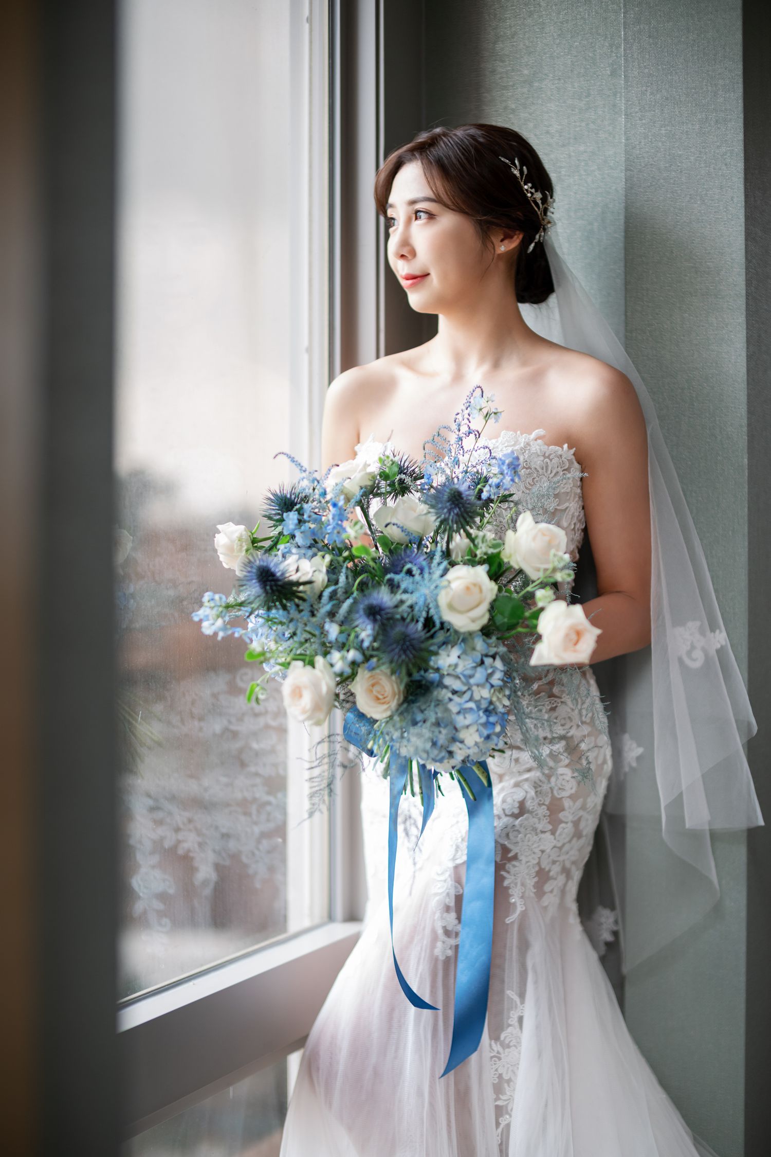 A bride in a lace wedding gown holds a stunning blue and white bouquet while gazing out a window in soft natural light.