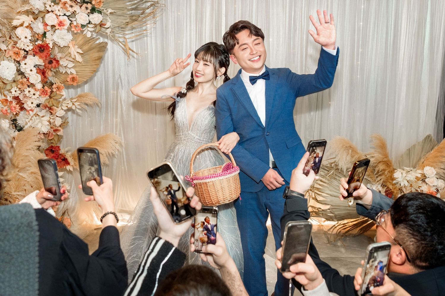 A couple waves to wedding guests taking photos with smartphones at a flower-decorated reception venue.