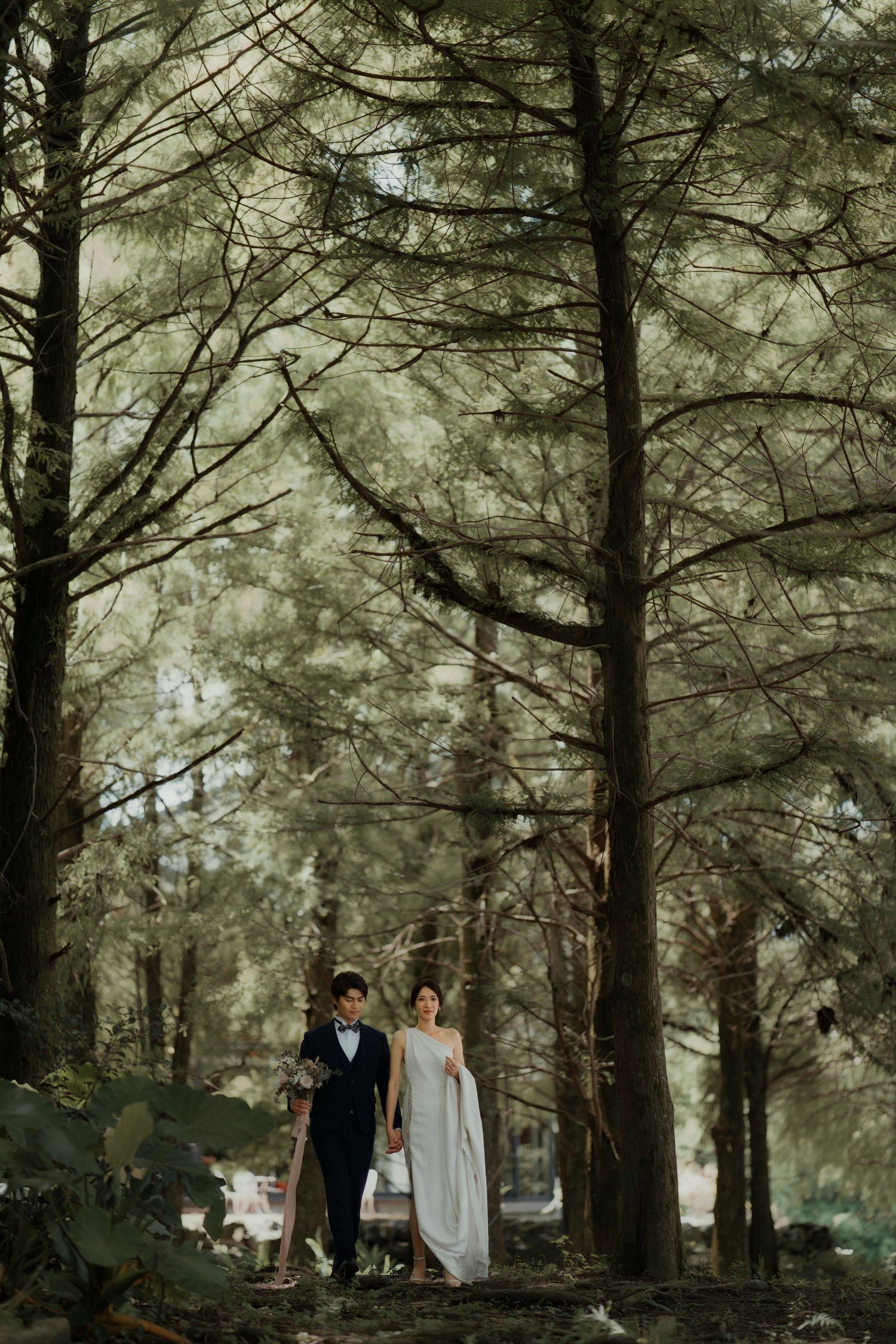 Couple walking through a moody pine forest with tall trees creating a dramatic, romantic atmosphere.