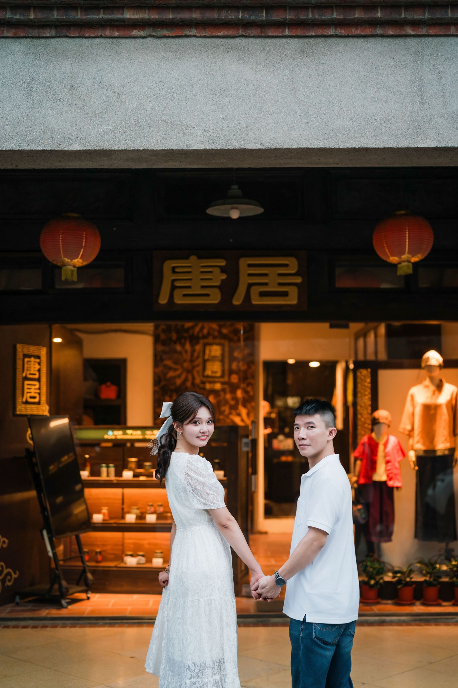 A couple in white outfits poses together at night in front of an Asian restaurant with red lanterns.