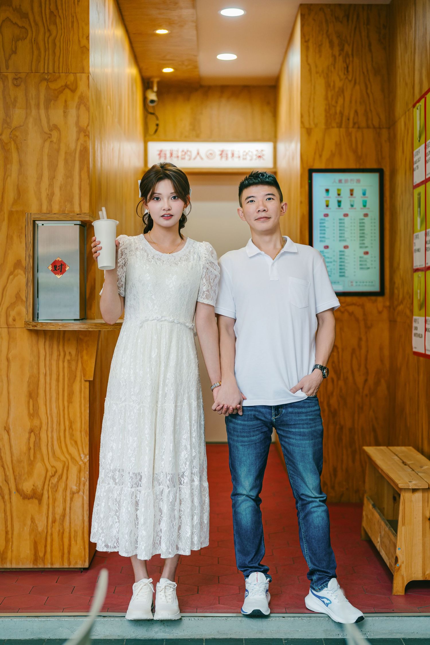 Two children stand together in a hallway wearing white outfits and jeans against a wooden paneled wall.
