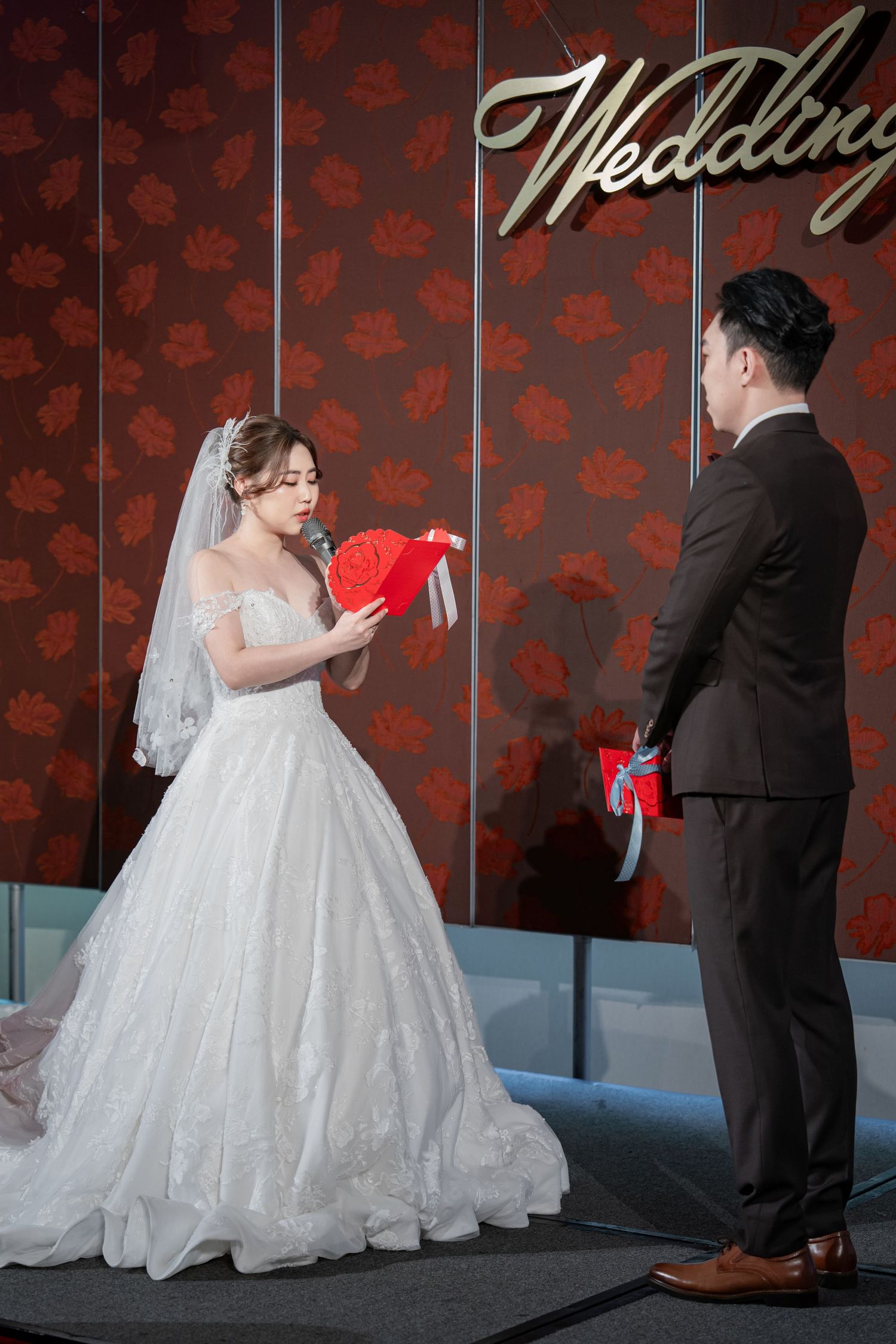A bride in a white ballgown and groom in dark suit share a playful moment against a red floral wallpaper backdrop.