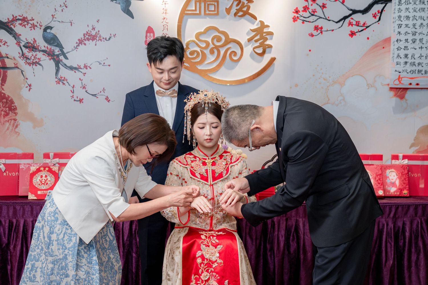 Bride in a red and gold Chinese wedding gown receives bracelets from an older couple during a traditional tea ceremony as a groom looks on.