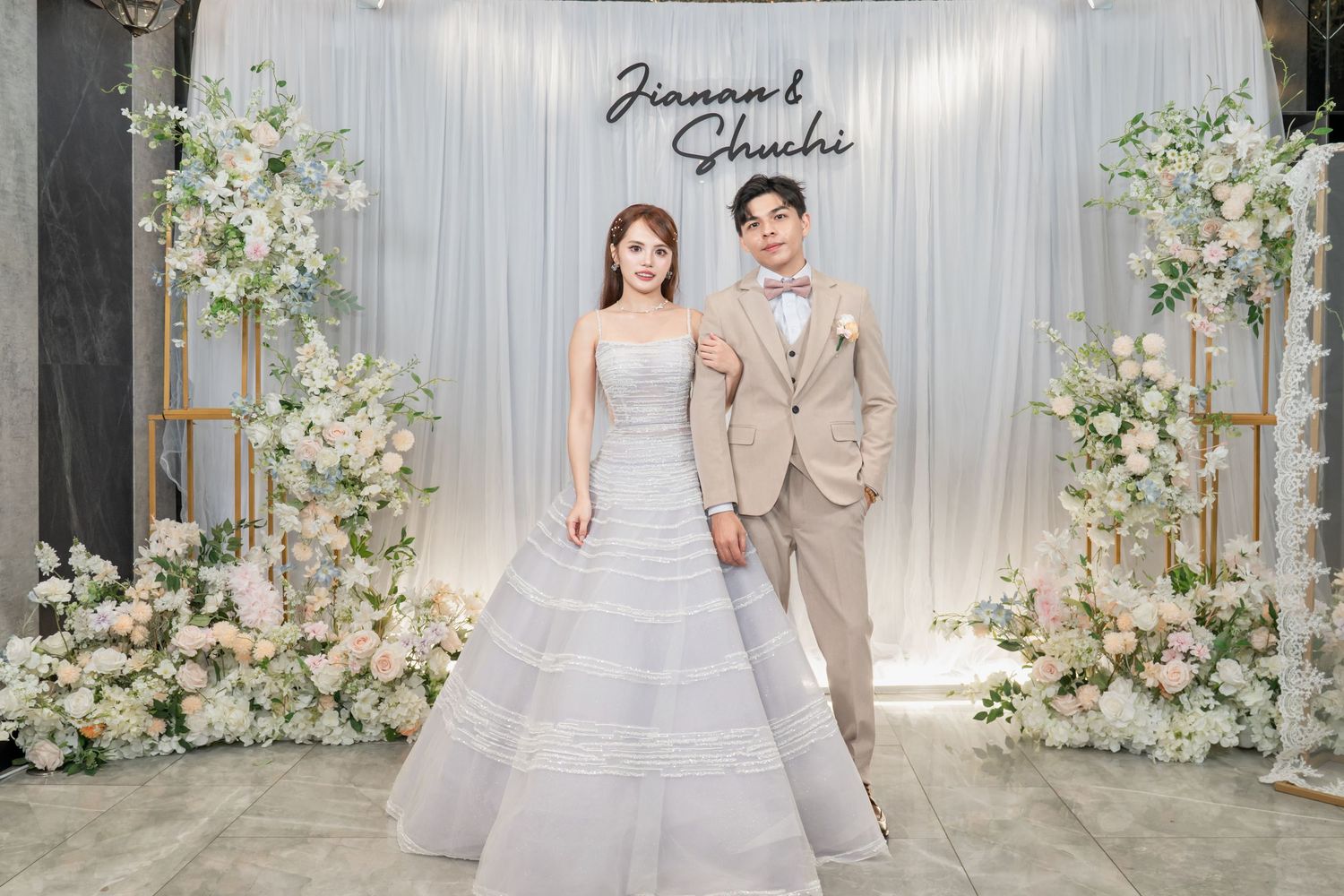 Bride in elegant gray ball gown and groom in beige suit pose together at a floral wedding reception backdrop.