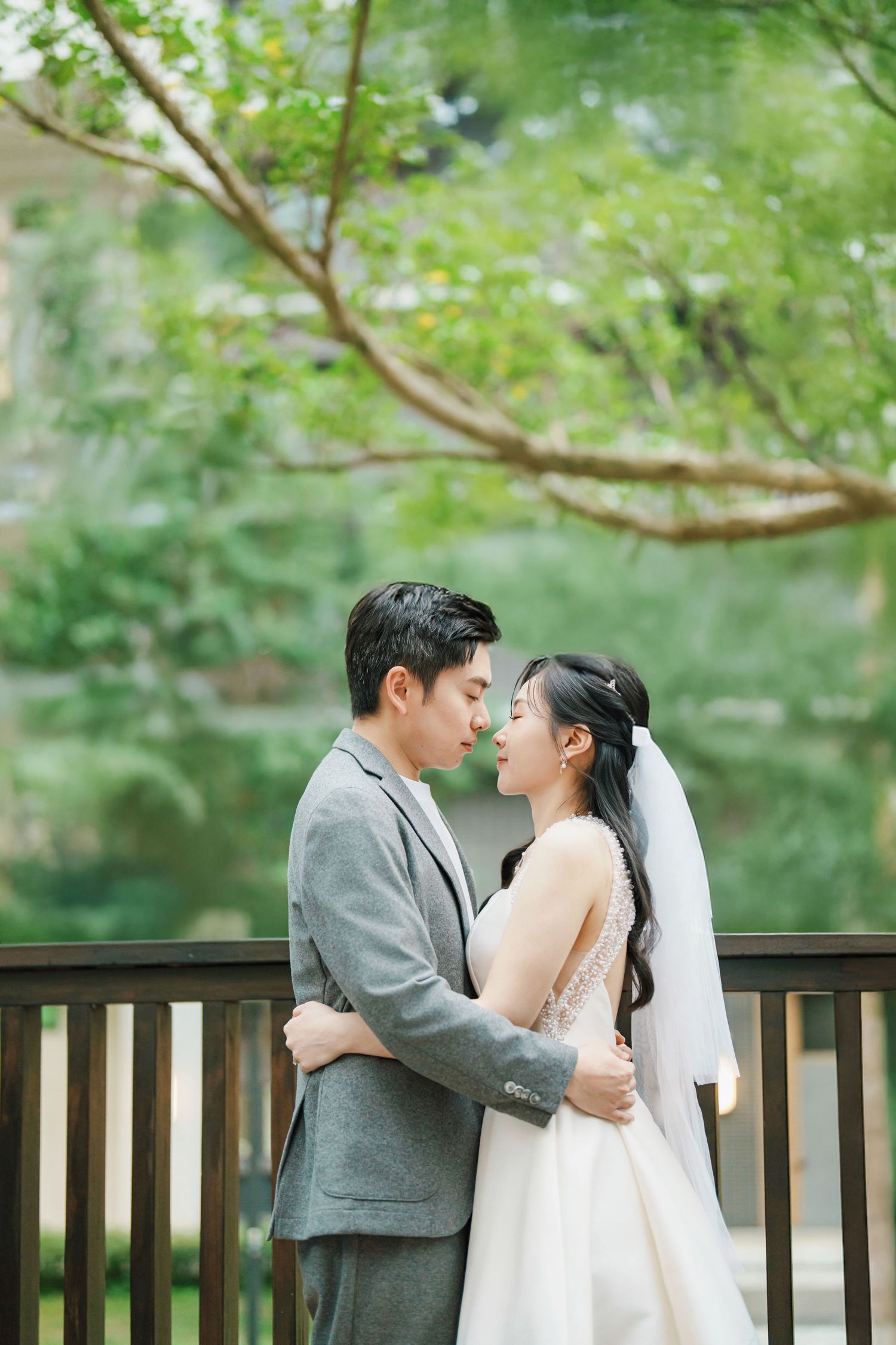 A romantic couple in wedding attire share intimate moments on a wooden deck surrounded by lush greenery.