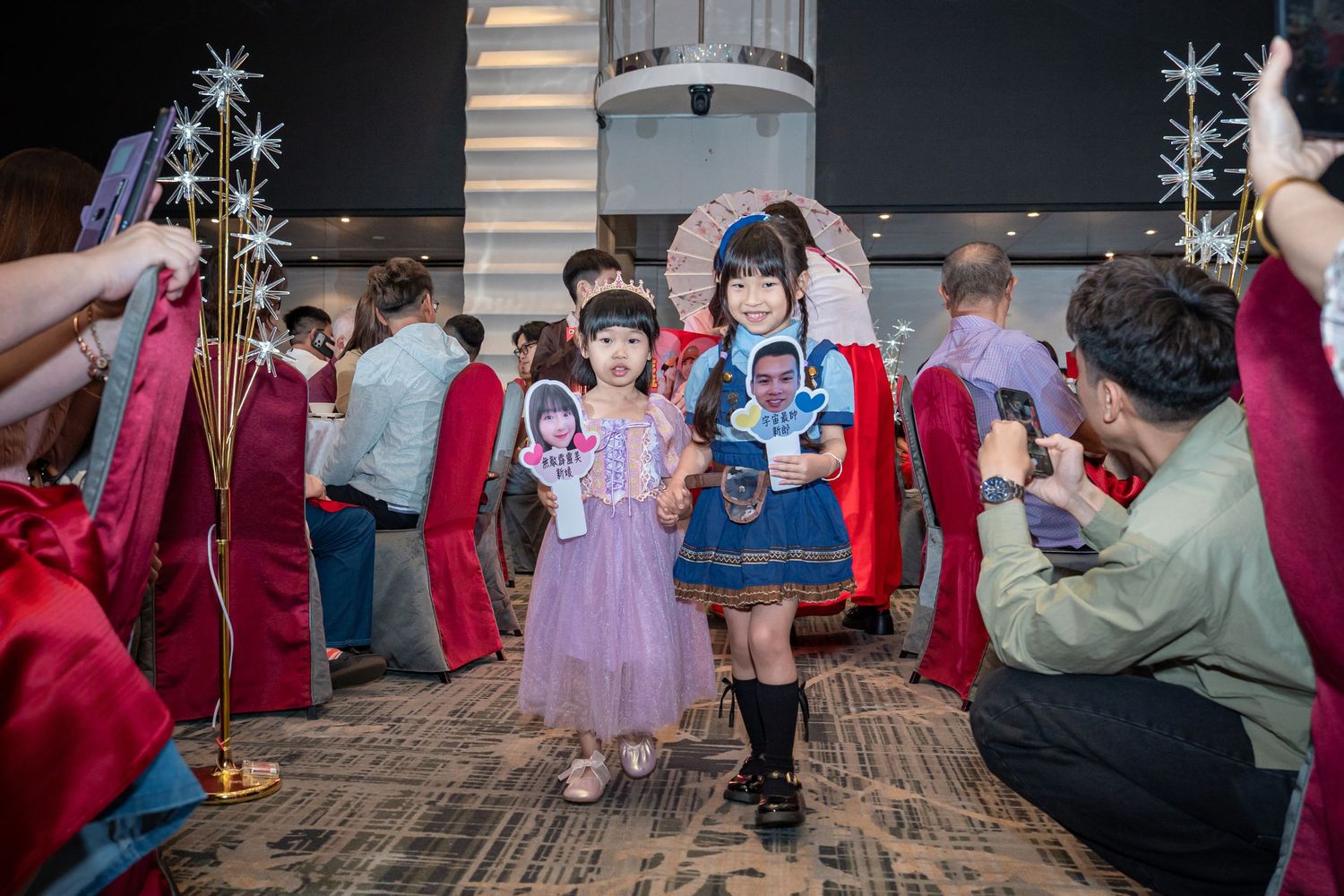 Two young girls in colorful dresses walk down an aisle at a festive event, holding decorative fans and parasols.
